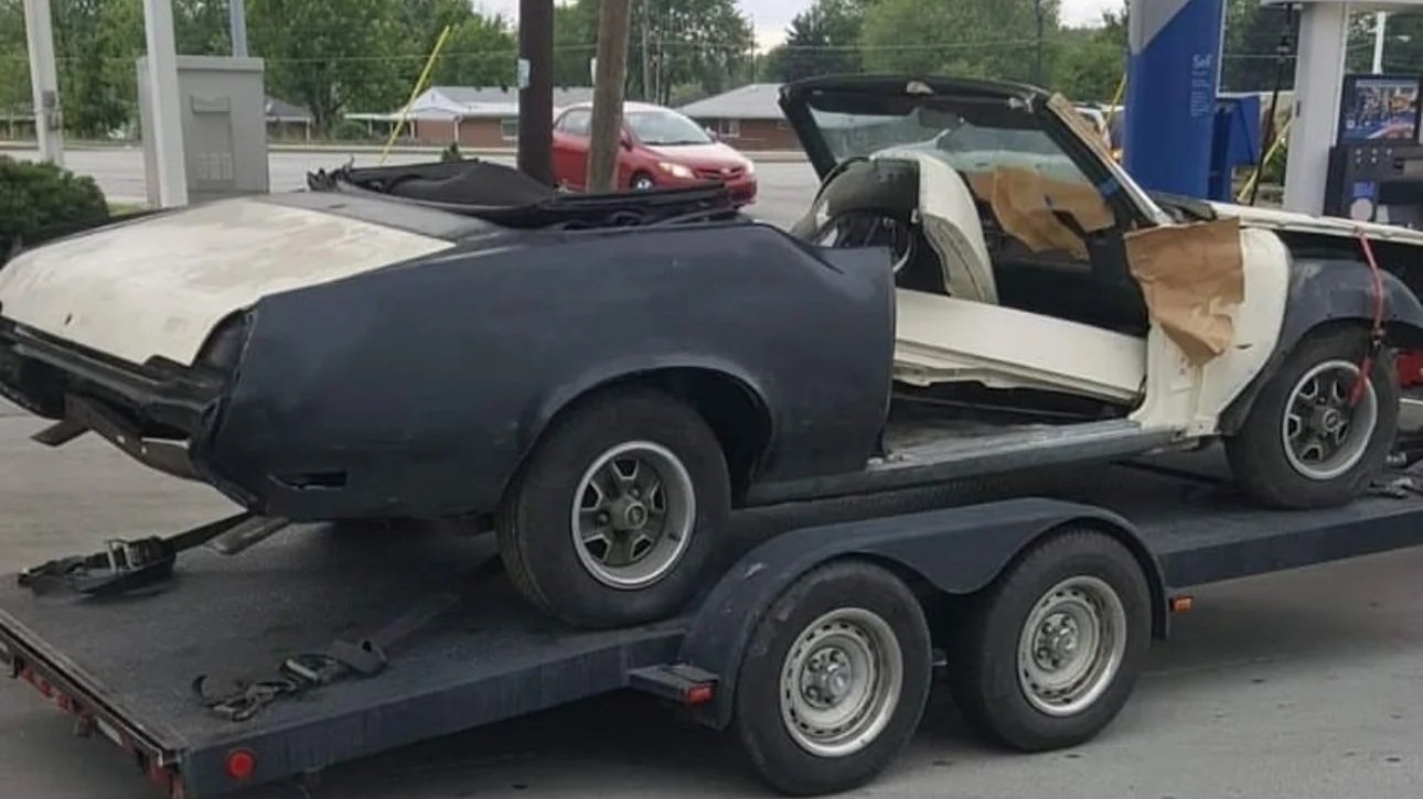 Before: a stripped Cutlass Supreme convertible in black primer and bare white panels, doors off, interior masked with paper, strapped down on a flatbed trailer