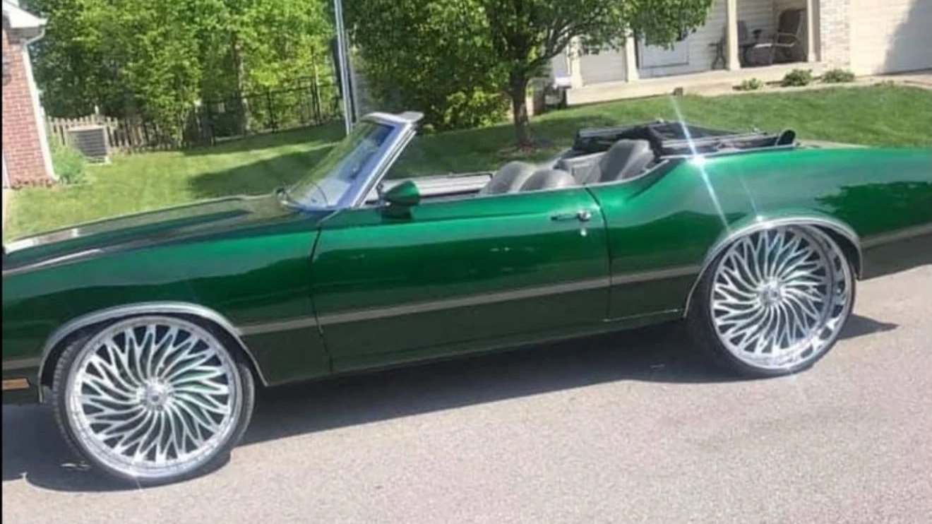 A Cutlass convertible finished in deep candy green with the top down, sitting on polished twisted-spoke wheels on a residential drive