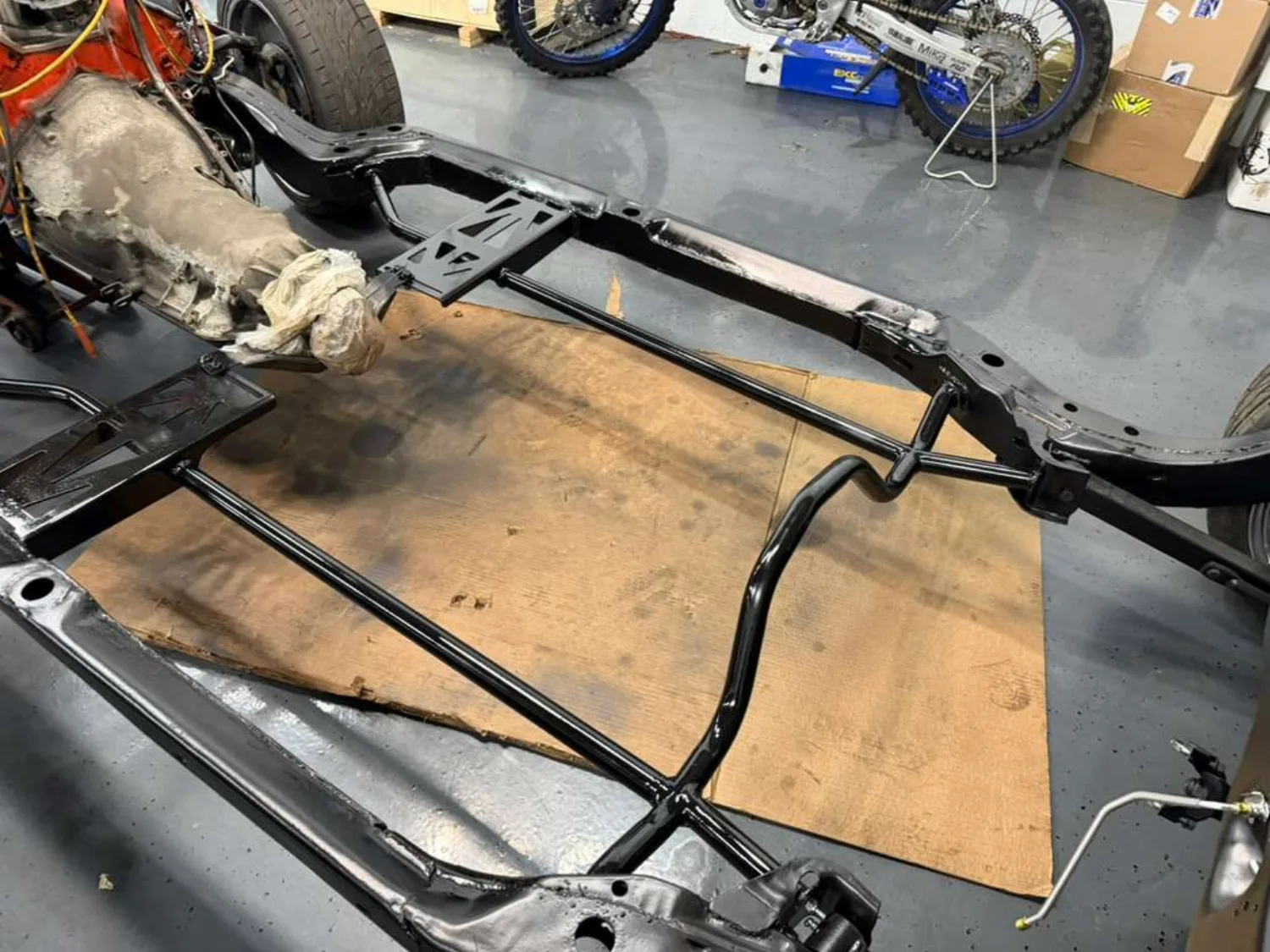A bare car frame on the shop floor with new fabricated bracing and gusset plates welded across the rails and crossmember, finished in black