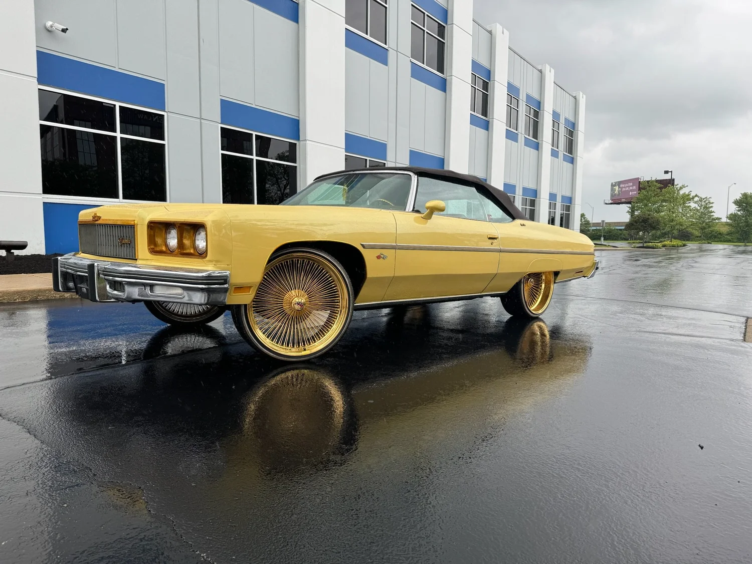 Yellow Chevrolet Caprice convertible on gold wire wheels, black top up, on wet pavement