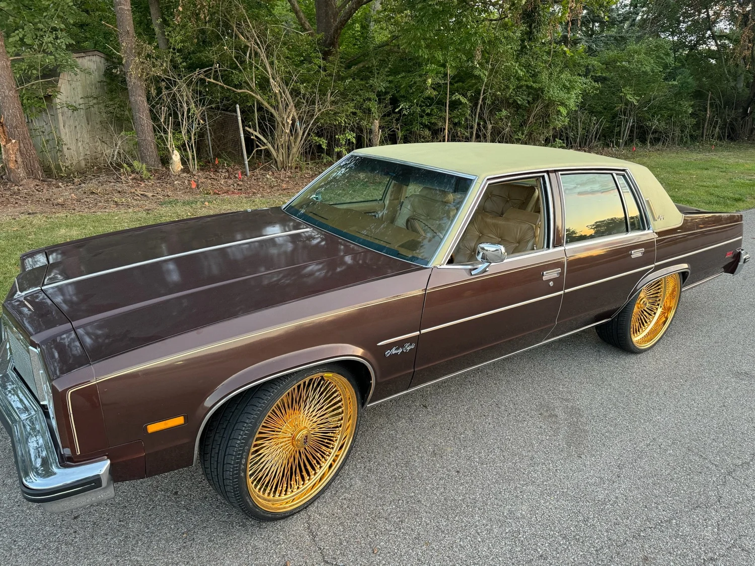 Brown Oldsmobile Ninety-Eight sedan with a tan vinyl top and gold wire wheels, parked at dusk