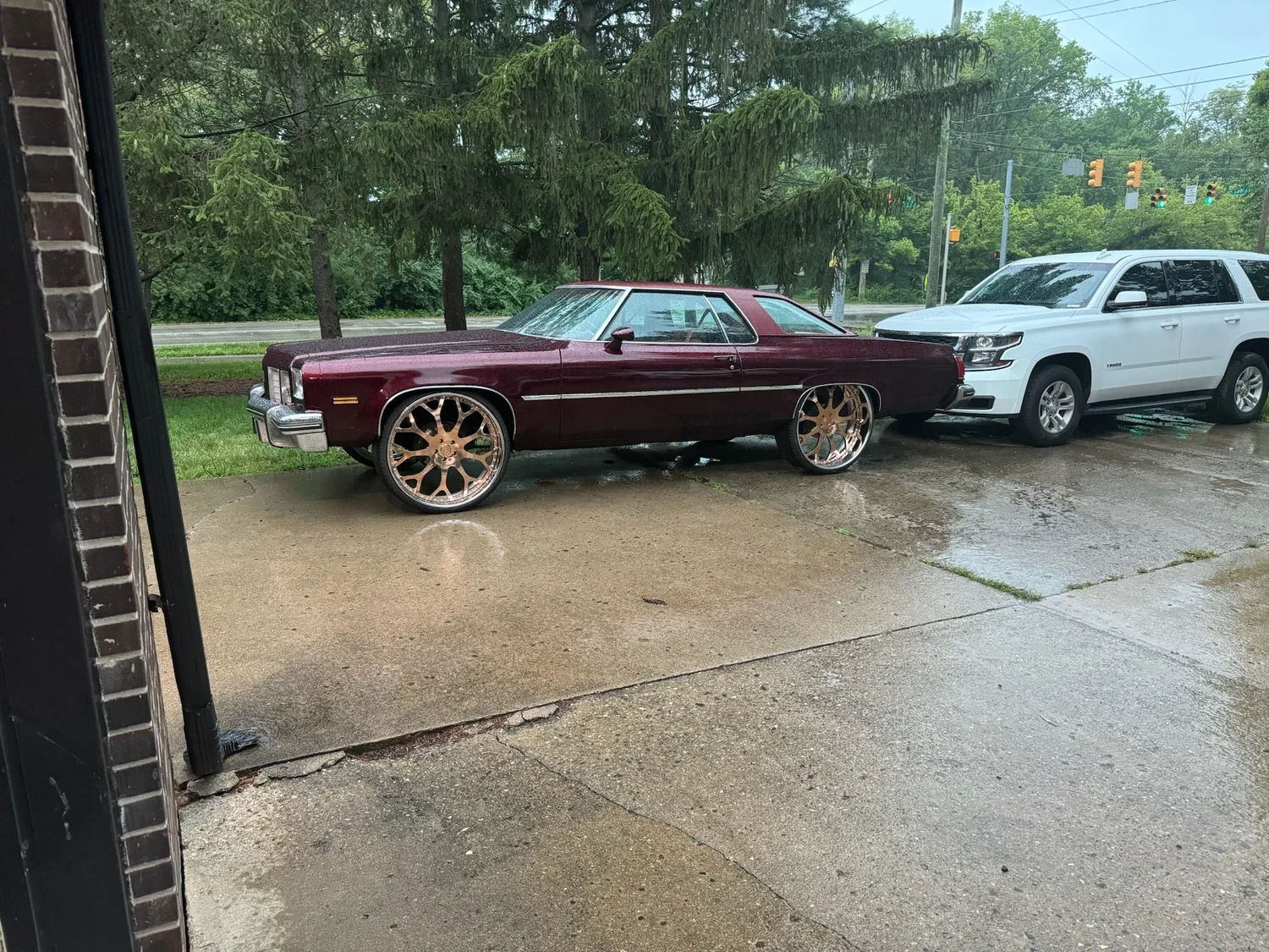 Burgundy metal-flake Cutlass Supreme on rose gold wheels, seen from inside the shop door on a rainy day