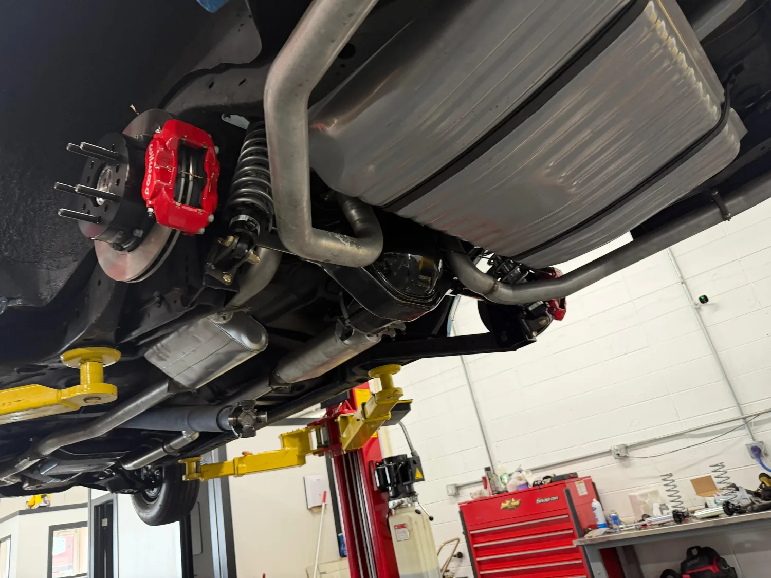 The full underside of a car on a two-post lift, showing the exhaust, driveshaft, rear axle and red brake calipers, the shop floor visible beyond